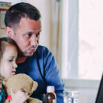 Father and child with teddy bear in front of laptop